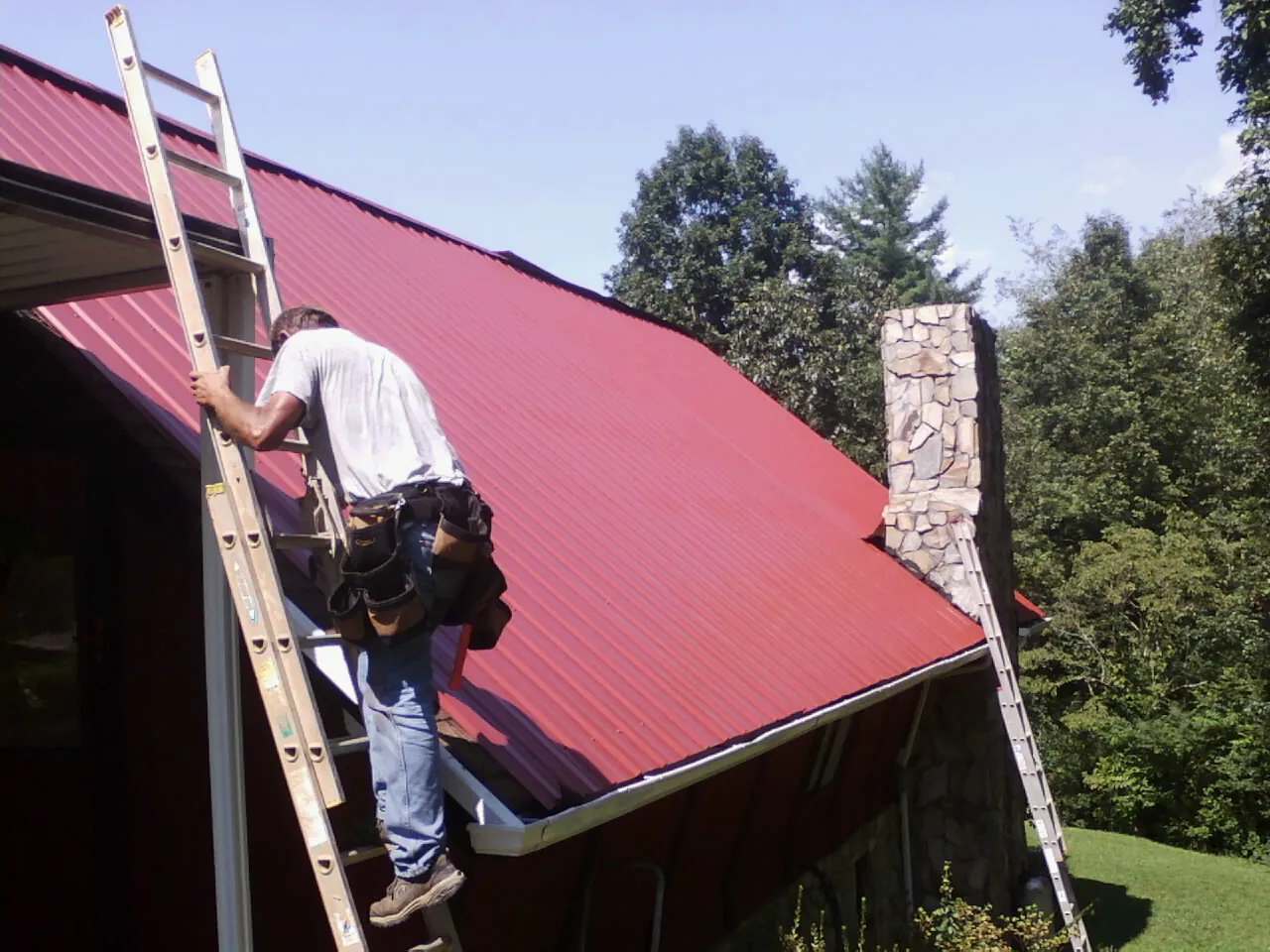 Roofer on a ladder beside a red metal roof and stone chimney — Soffit & Fascia Repair by Roofing Brothers in New Church